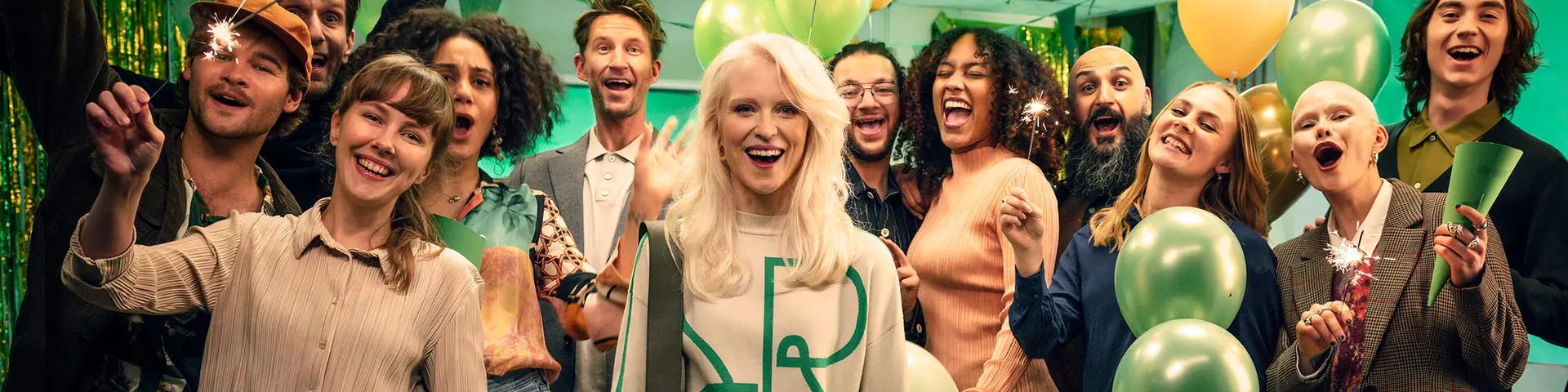 En stor samling människor står i ett rum fyllt med girlanger, ballonger, flaggspel och mer. Alla ler och har kul på bilden.