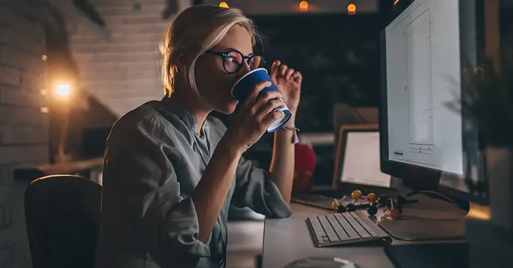 Ny på jobbet? Ung kvinna dricker kaffe framför datorn. 