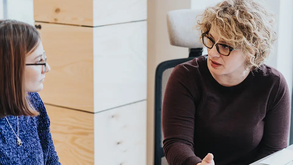 Två kvinnliga kollegor pratar vid ett skrivbord. 