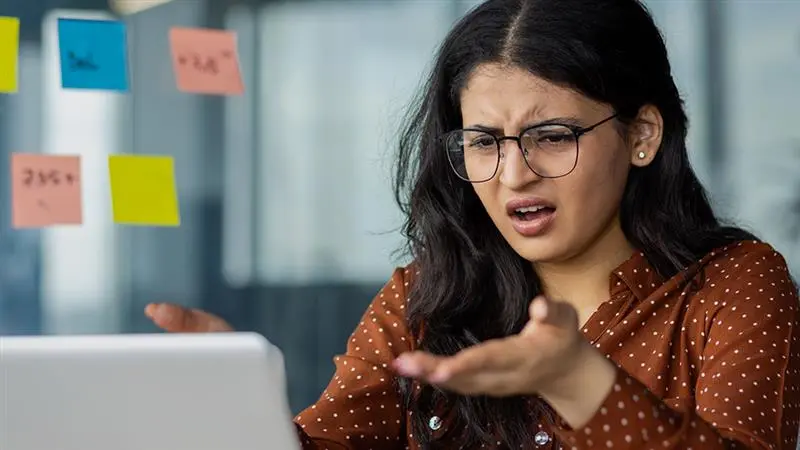 Kvinna i brun skjorta är arg på en dator som trasslar. 