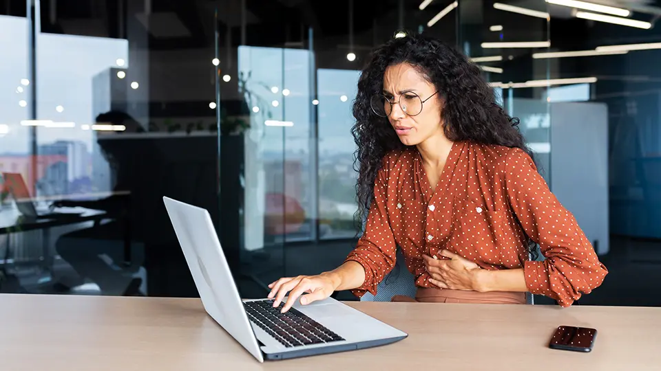 Kvinna vid laptop håller sig för magen och har ont. 