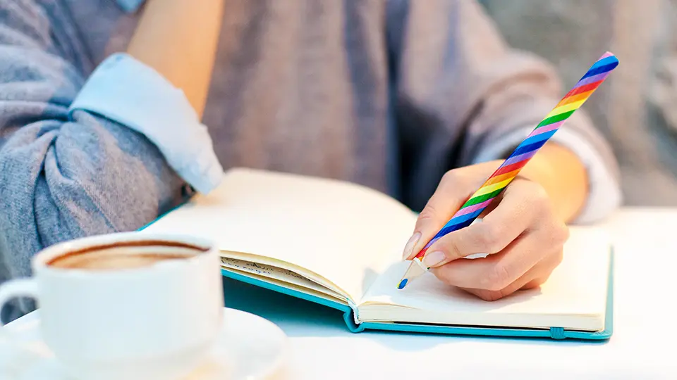 Kvinna med pride-färgad penna skriver i skrivbok. 