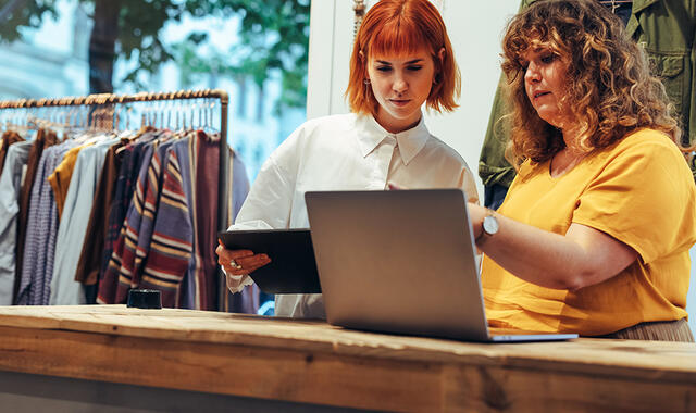 Två kvinnor, som jobbar i butik, står i kassan och kollar på datorn. 