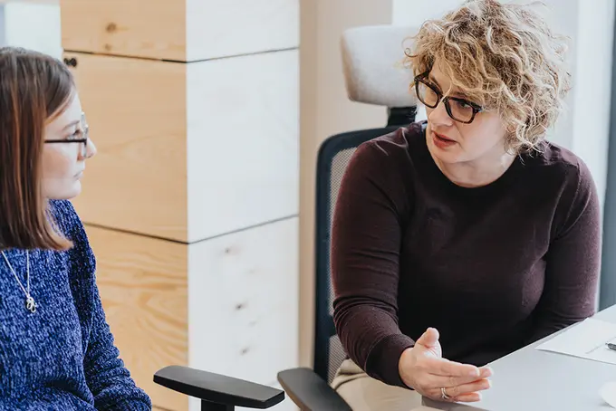 Två kvinnliga kollegor pratar vid ett skrivbord. 