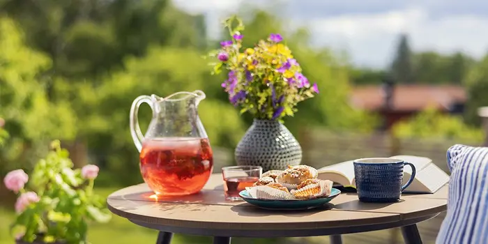 Somrigt frukostbord utomhus med saft, kaffe, bakverk och bok – symbol för semester och avkoppling.